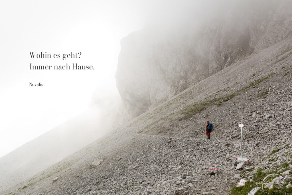 Einsamer Wanderer in einer nebligen Fels-/Gerölllandschaft mit einem Zitat von Novalis "Wohin es geht? Immer nach Hause.". Kontemplative Fotografie aus dem Projekt "Zwischen Tau und Wind" von Katrin Jeschke