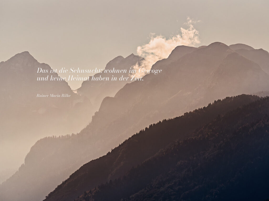 Bergkette im Dunst mit einer einzelnen Wolke und einem Text von Rilke: "Das ist die Sehnsucht: wohnen im Gewoge und keine Heimat haben in der Zeit." Kontemplative Fotografie aus dem Projekt "Zwischen Tau und Wind" von Katrin Jeschke