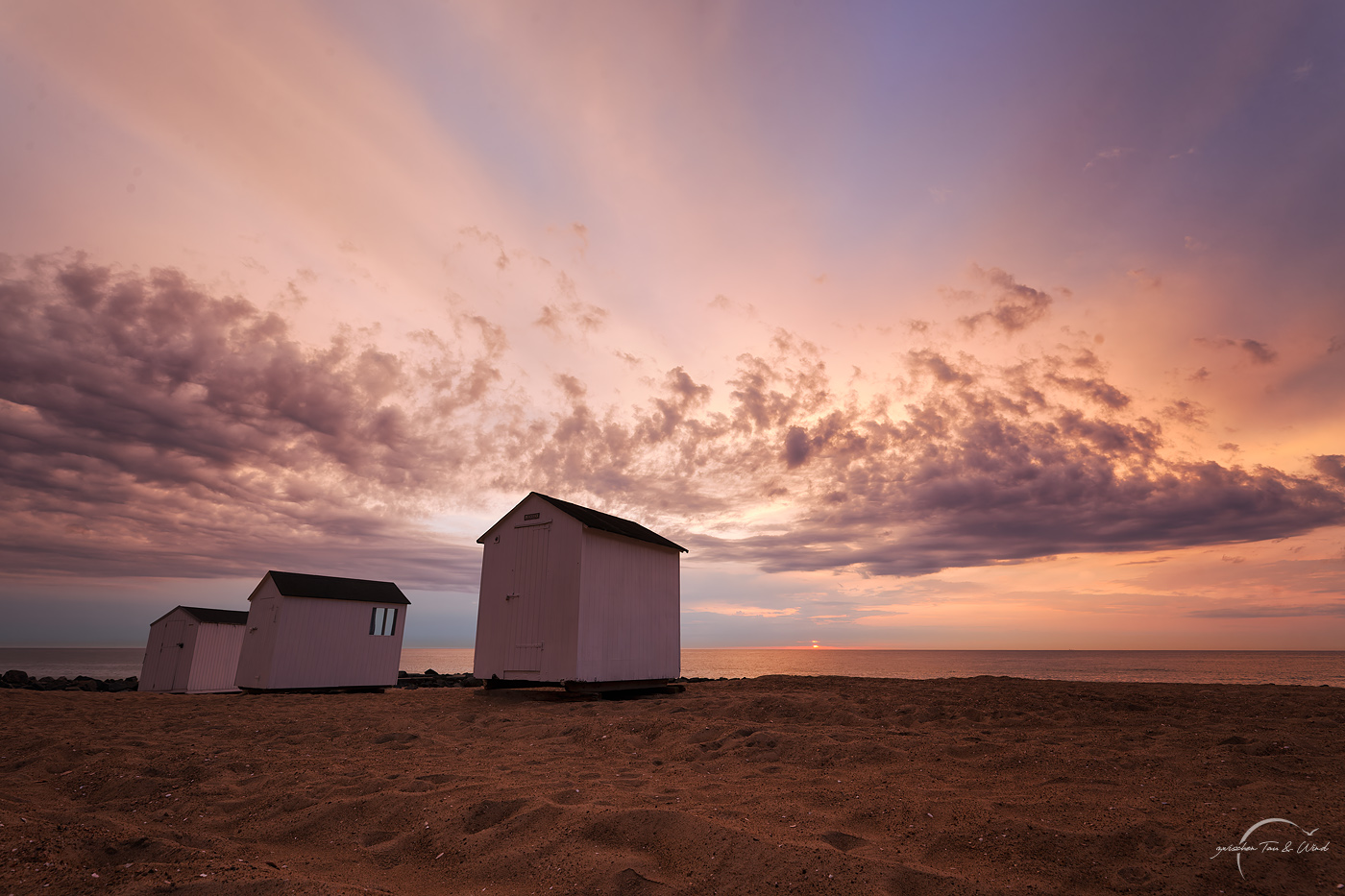 Drei kleine, weiße Häusschen am Nørlev Strand in Dänemark. Im Hintergrund geht kaum sichtbar gerade die Sonne unter. Der Himmel ist von ein paar Wolken durchzogen und in orange-gelb-magenta-blaue Farben getaucht. Kontemplative Fotografie aus dem Projekt "Zwischen Tau und Wind" von Katrin Jeschke