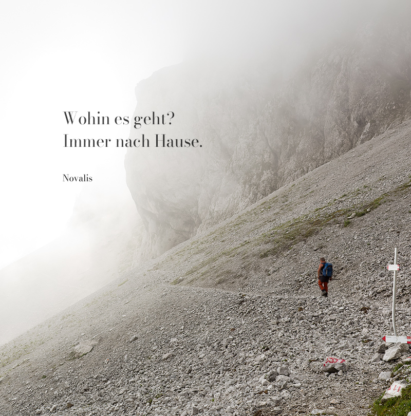 Einsamer Wanderer in einer nebligen Fels-/Gerölllandschaft mit einem Zitat von Novalis "Wohin es geht? Immer nach Hause.". Kontemplative Fotografie aus dem Projekt "Zwischen Tau und Wind" von Katrin Jeschke