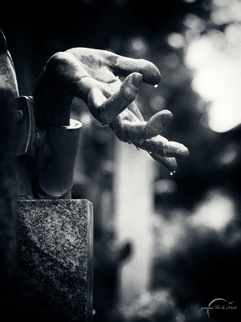 Schwarz-weiß-Nahaufnahme einer steinernen Handskulptur nach dem Regen. Die Hand ist herabhängend, ihre Finger sind leicht gekrümmt, und kleine Wassertropfen perlen von den Fingerspitzen ab. Kontemplative Fotografie aus dem Projekt "Zwischen Tau und Wind" von Katrin Jeschke